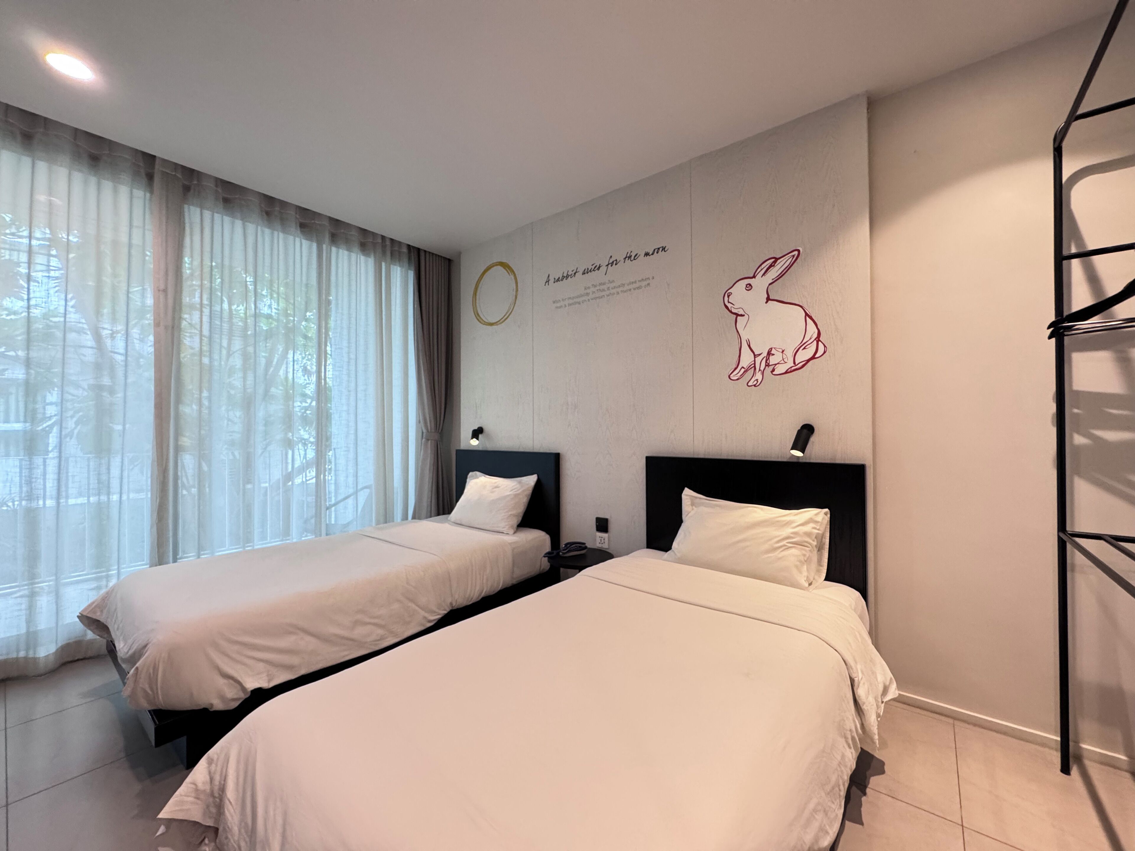 Habitación de hotel con dos camas, una ventana con cortinas translúcidas, una lámpara de pared y una ilustración decorativa de un conejo.
