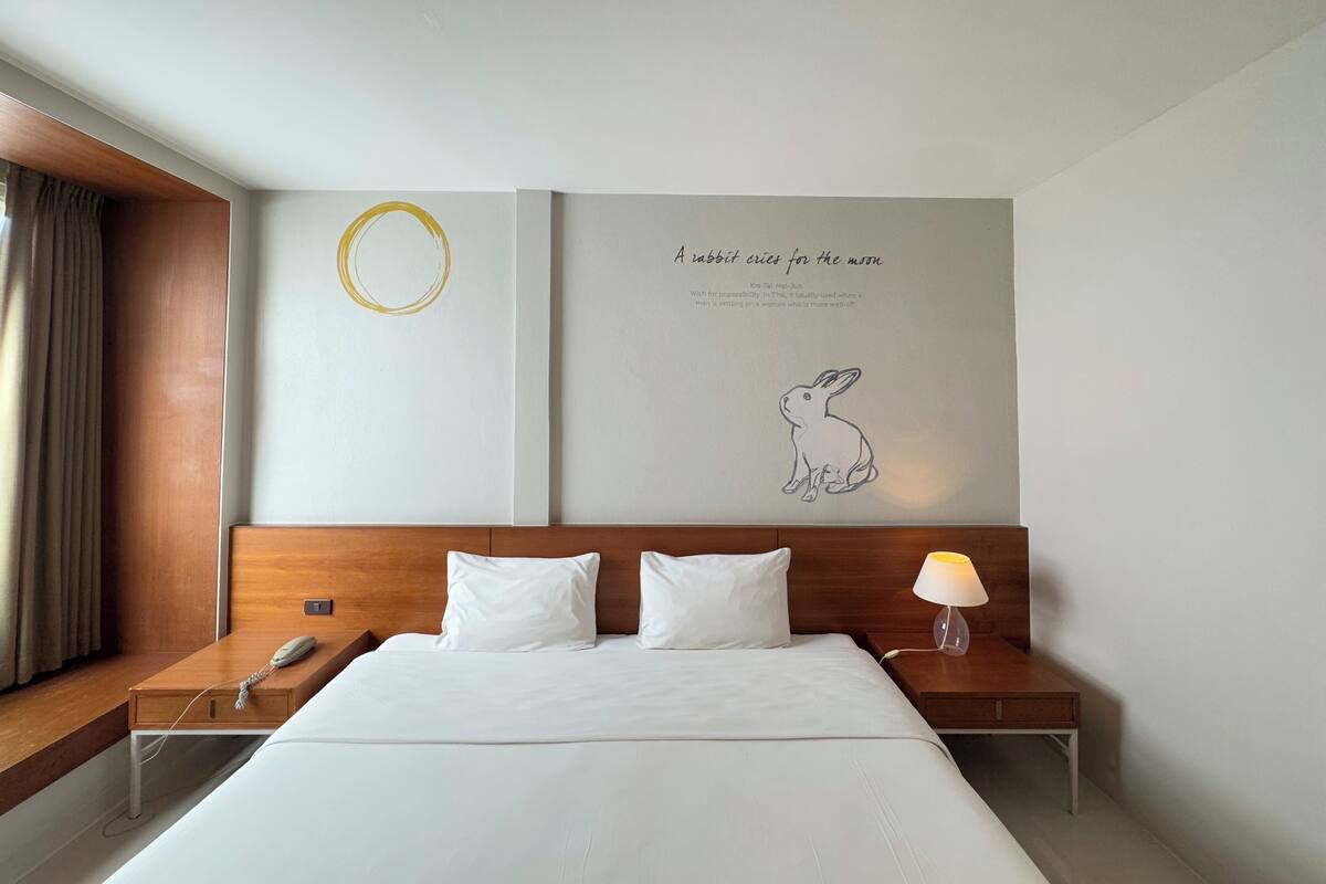 A hotel room with a large bed, a wooden headboard, a bedside table with a lamp, and a wall with a circular design and a rabbit illustration.