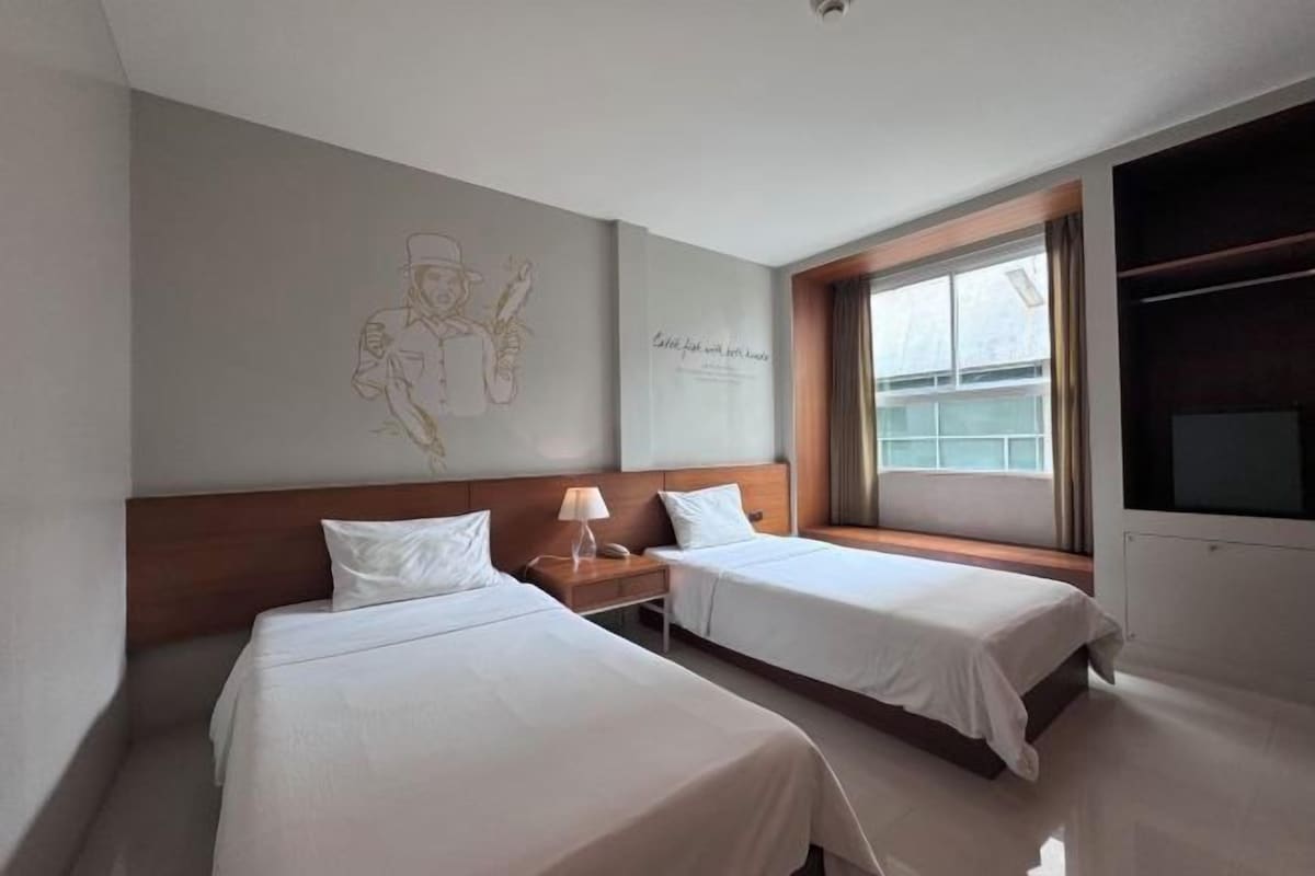 A hotel room with two single beds, a nightstand with a lamp, a large window, and a wall with a decorative sketch.