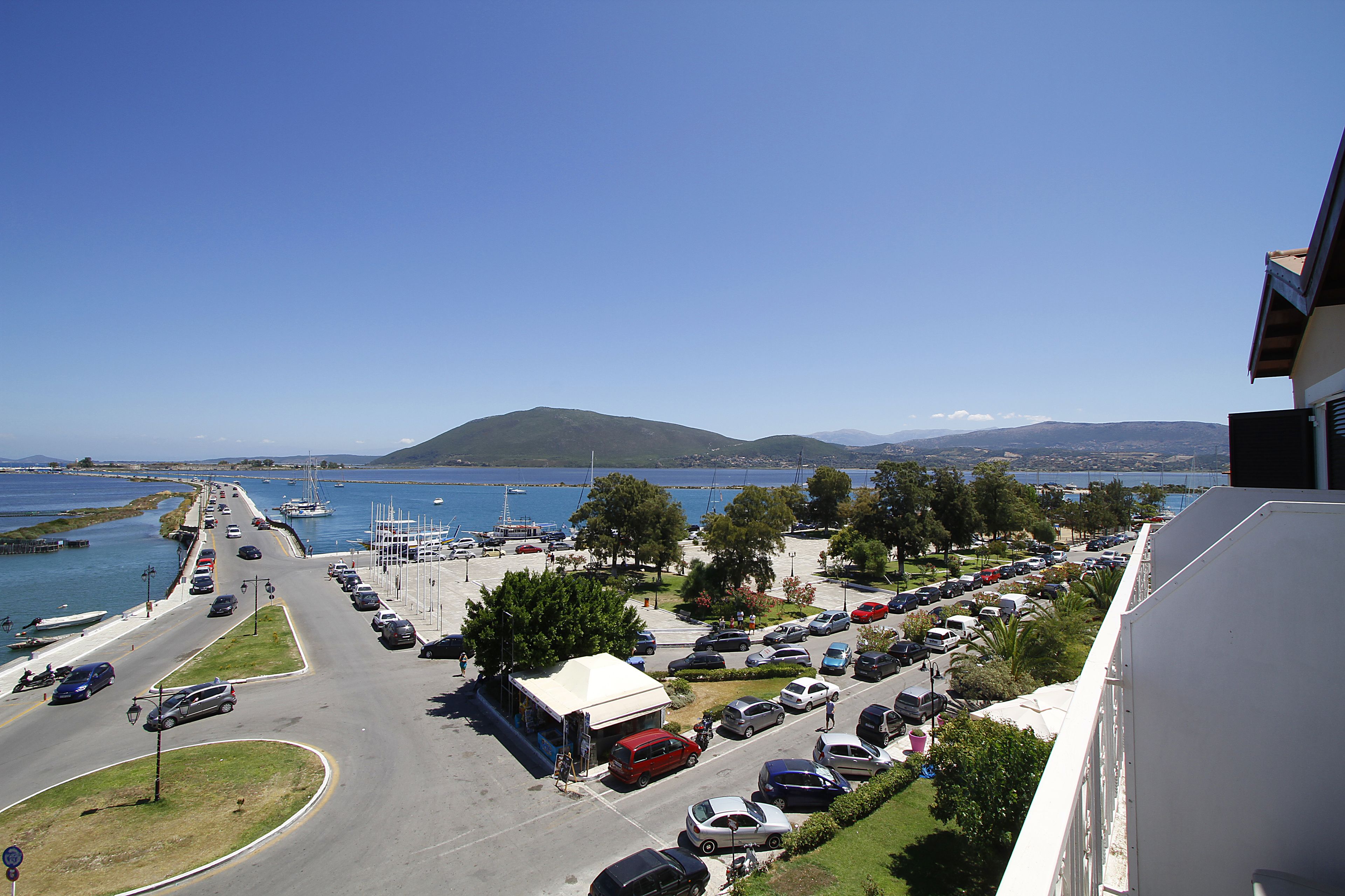 A coastal road with parked cars, a white building with a white roof, and a view of the sea and hills in the background.