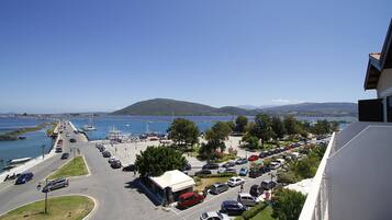 A coastal road with parked cars, a white building with a white roof, and a view of the sea and hills in the background.