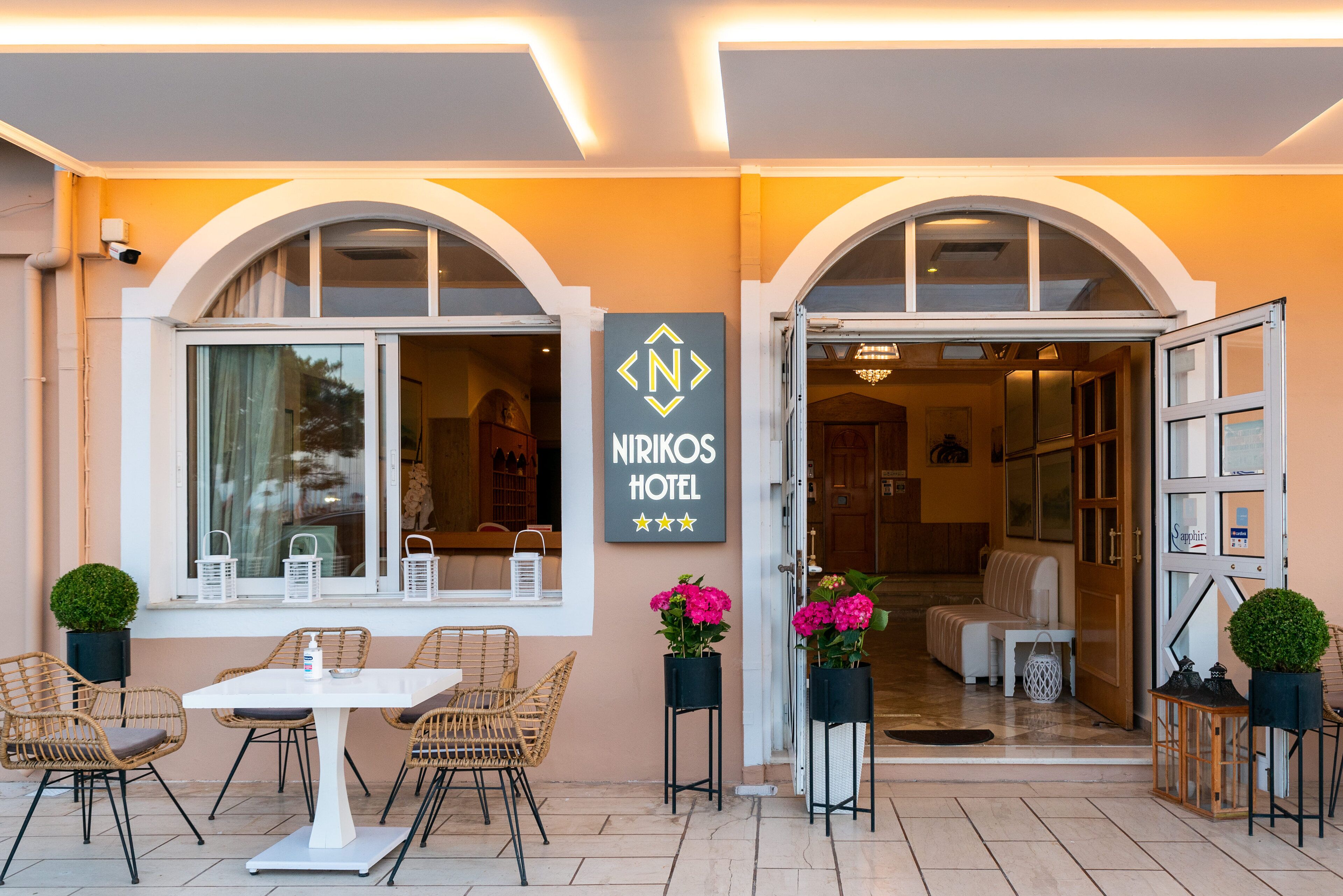 L’entrée d’un hôtel, avec une enseigne indiquant « HÔTEL NIKOS », et un espace détente comprenant des fauteuils en osier et une table.
