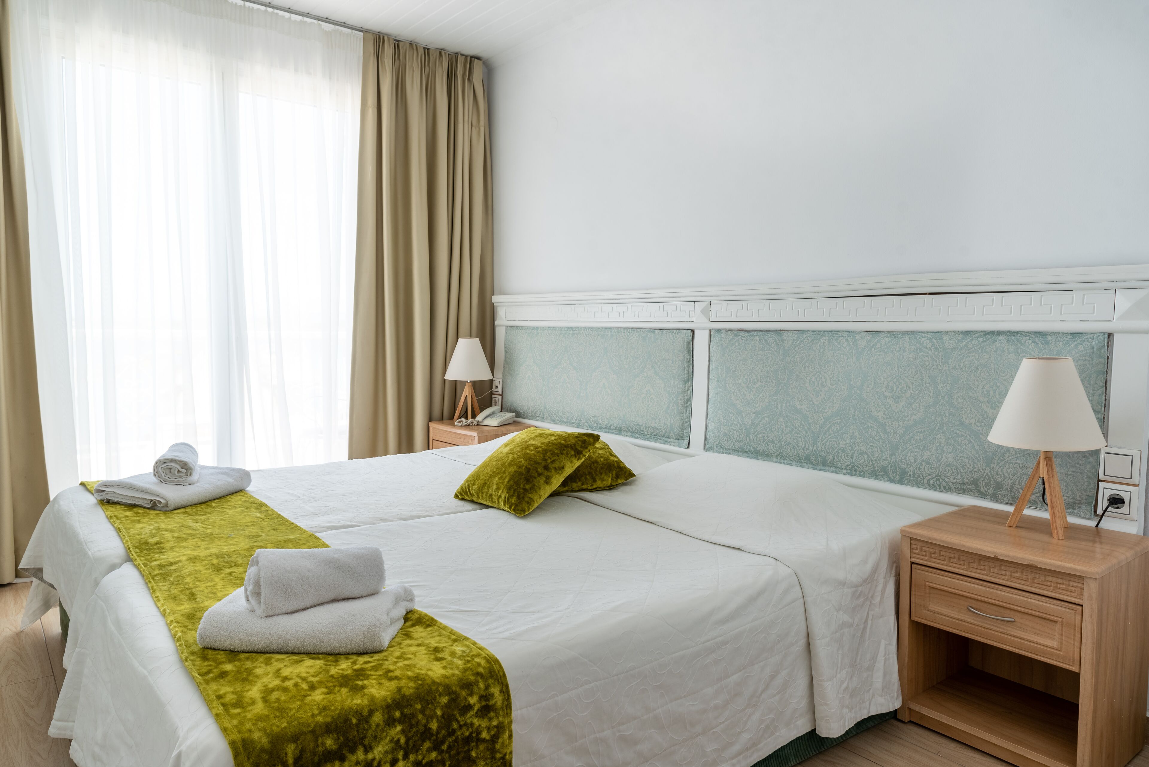 A hotel room with two beds, a nightstand with a lamp, a window with curtains, and a wall with a patterned design.