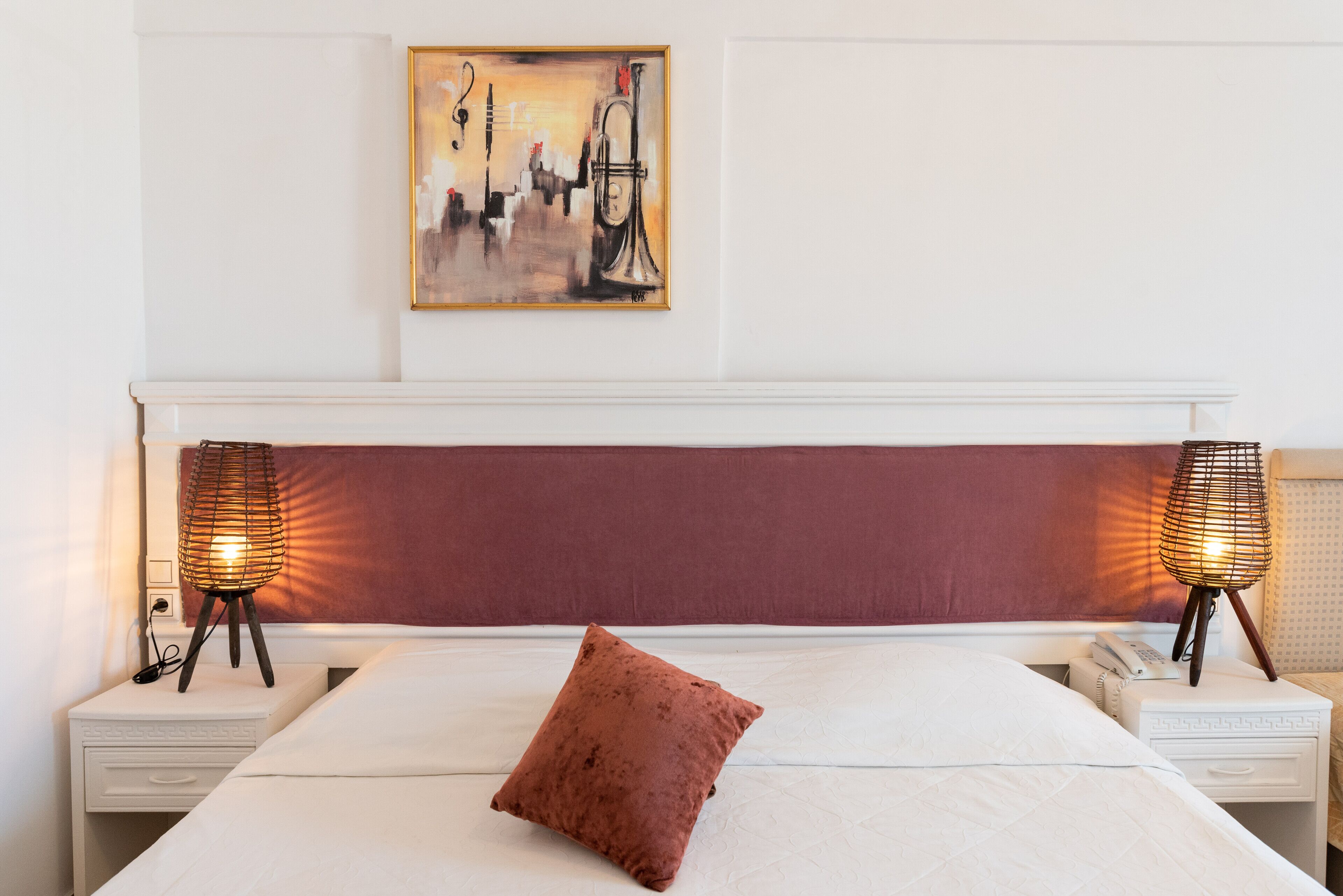 Un lit bien fait, avec un seul oreiller marron, deux lampes de chevet, une table de chevet blanche avec un téléphone, et un tableau abstrait encadré accroché au mur.
