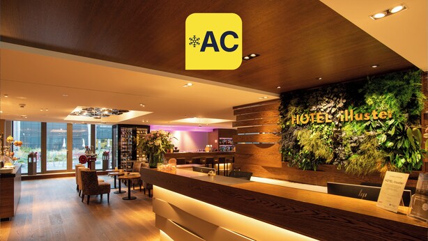 Zona de recepción de un hotel con un mostrador de madera, una pared verde con plantas y vistas a la ciudad a través de amplias ventanas.