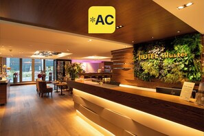 A hotel reception area with a wooden reception desk, a green wall with plants, and a view of the city through large windows. - HOTEL illuster - Urban & Local (Uster)