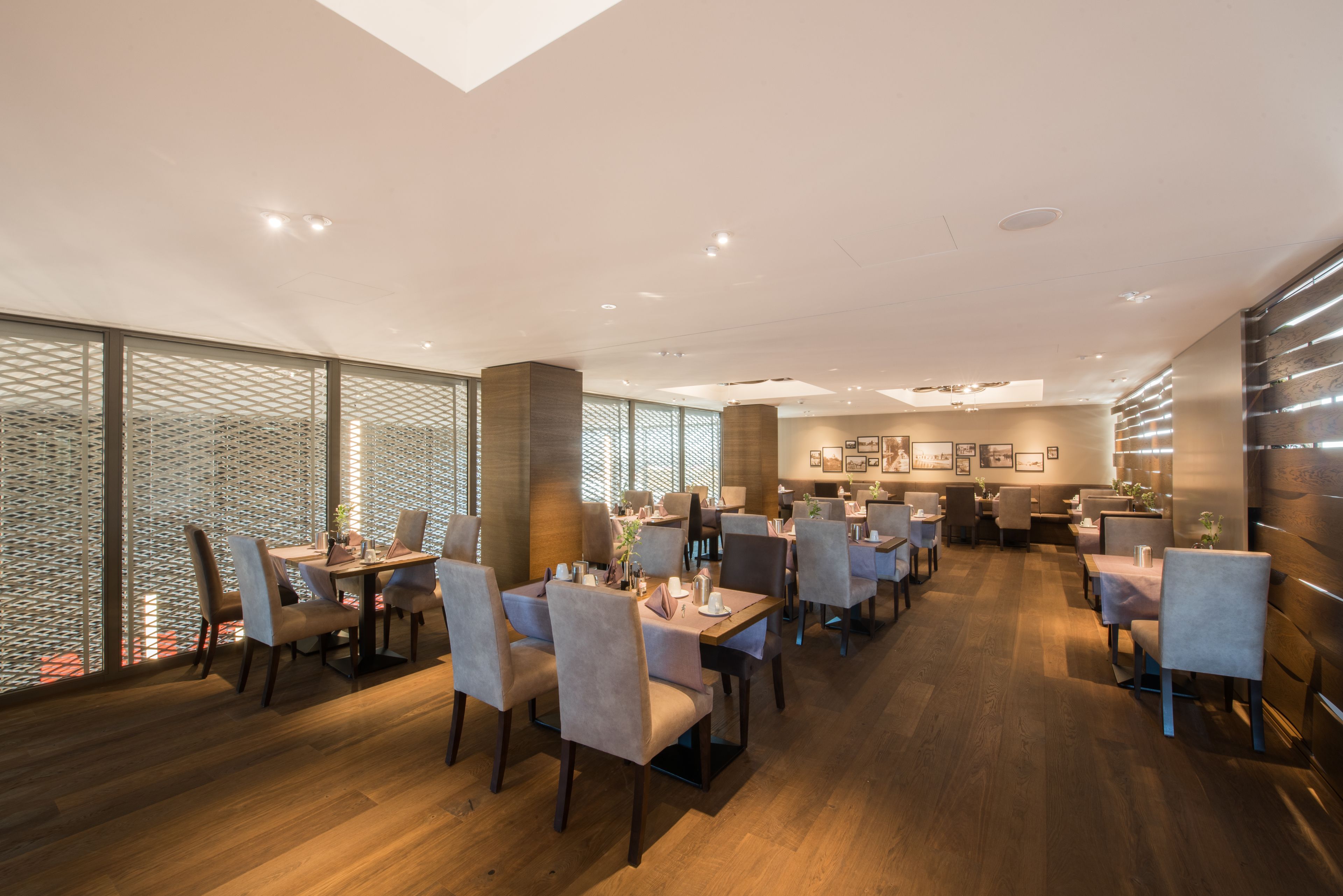 A modern restaurant with wooden flooring, multiple dining tables with chairs, and large windows with a view.