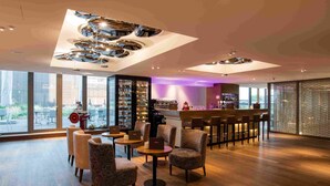 A modern lounge area with wooden flooring, comfortable seating, a bar with stools, and a large window offering a view of the outdoors. - HOTEL illuster - Urban & Local (Uster)