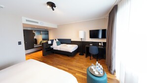 A modern hotel room with a large bed, a desk, a flat-screen TV, and a seating area with a sofa and a chair. - HOTEL illuster - Urban & Local (Uster)