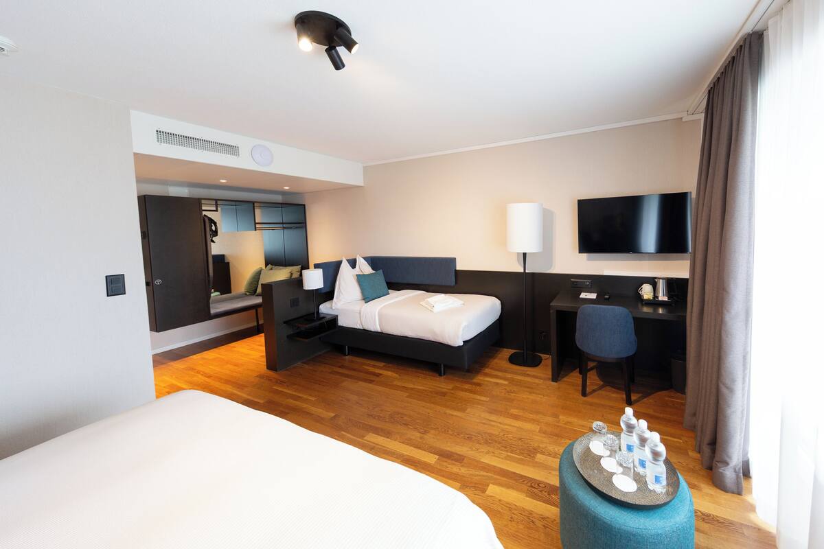 A modern hotel room with a large bed, a desk, a flat-screen TV, and a seating area with a sofa and a chair.