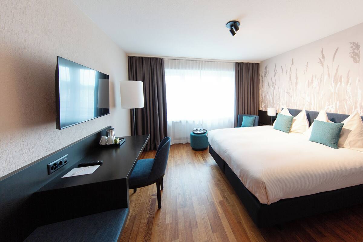 A modern hotel room with a large bed, a desk with a flat-screen TV, a coffee maker, and a small round table.