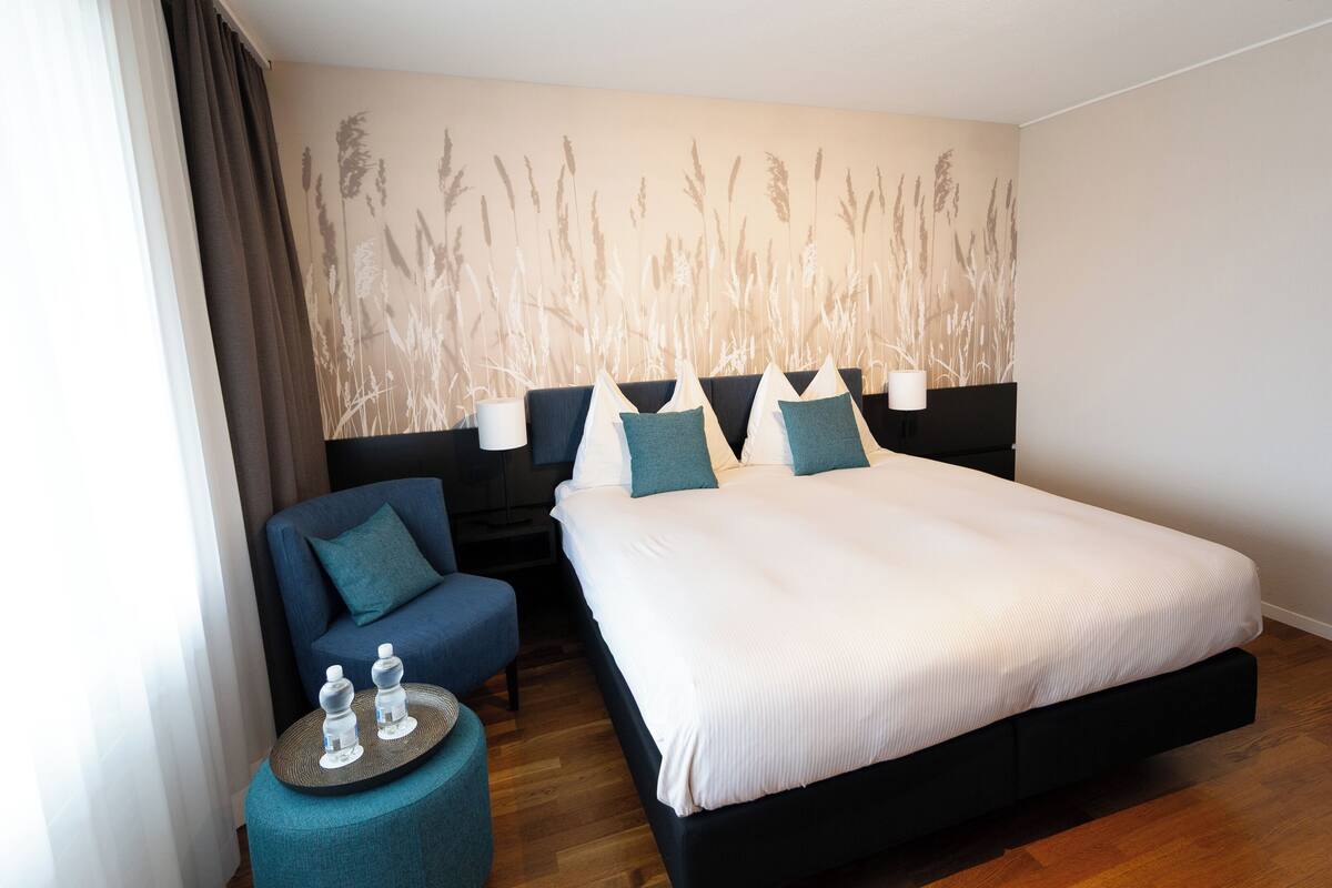 A hotel room with a large bed, a bedside table with water bottles, a blue armchair, and a wall with a nature-themed mural.