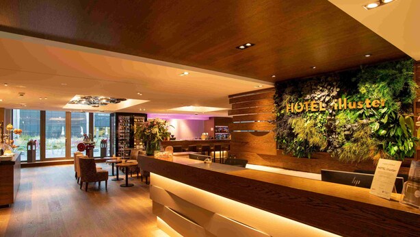 A hotel reception area with a wooden reception desk, a wall-mounted plant display, and a view of the city through large windows. - HOTEL illuster - Urban & Local (Uster)
