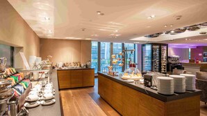 A modern hotel lobby with a central reception desk, a buffet area with various food items, and a seating area with chairs. - HOTEL illuster - Urban & Local (Uster)