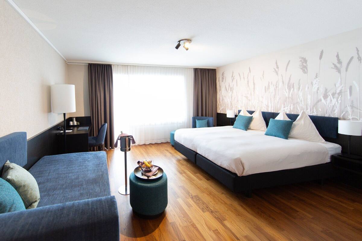 A modern hotel room with a large bed, a sofa, a desk, and a small round ottoman.