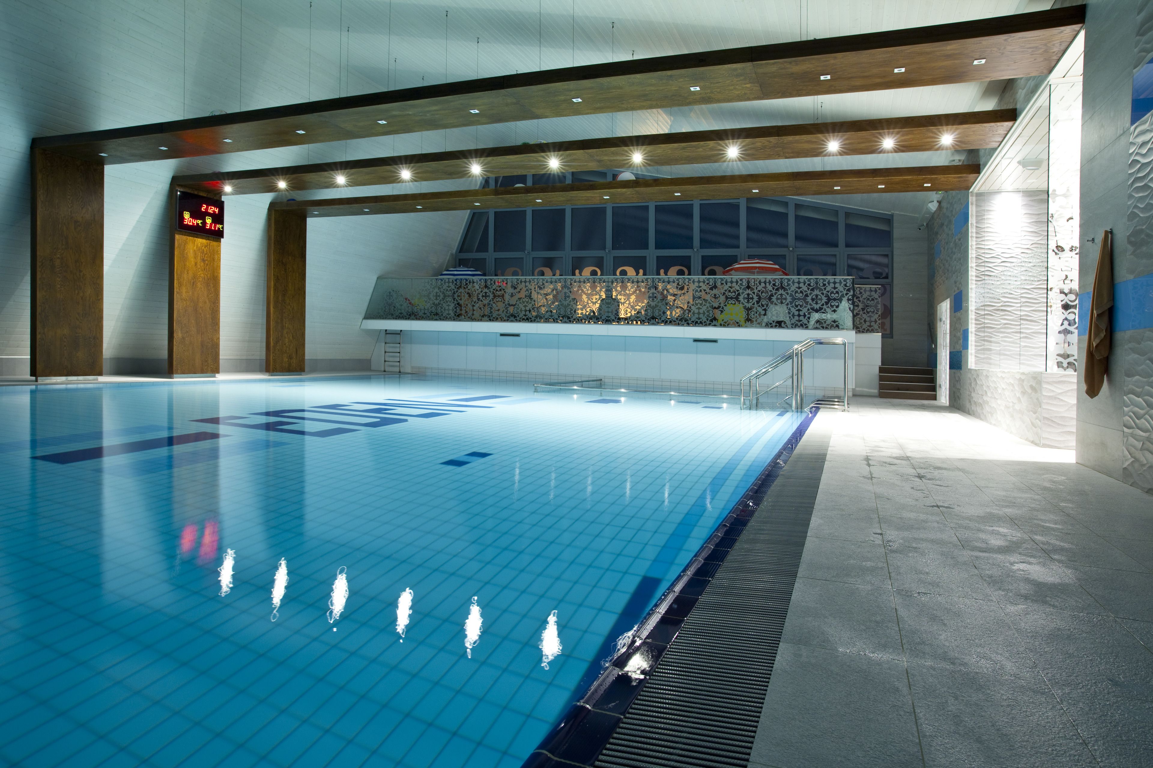 A large indoor swimming pool with a tiled floor, a wooden ceiling, and a digital clock displaying the time.