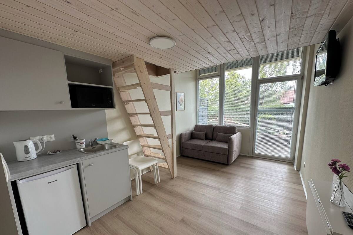 Una cocina moderna con microondas integrado, cafetera y refrigerador pequeño. Se ve una escalera de madera apoyada contra la pared, un sofá gris y una ventana con persianas.