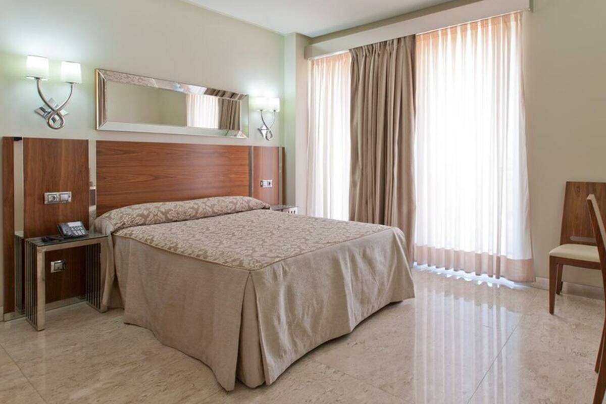 Une chambre d’hôtel avec un grand lit, un bureau avec une chaise, un miroir et une fenêtre avec des rideaux.