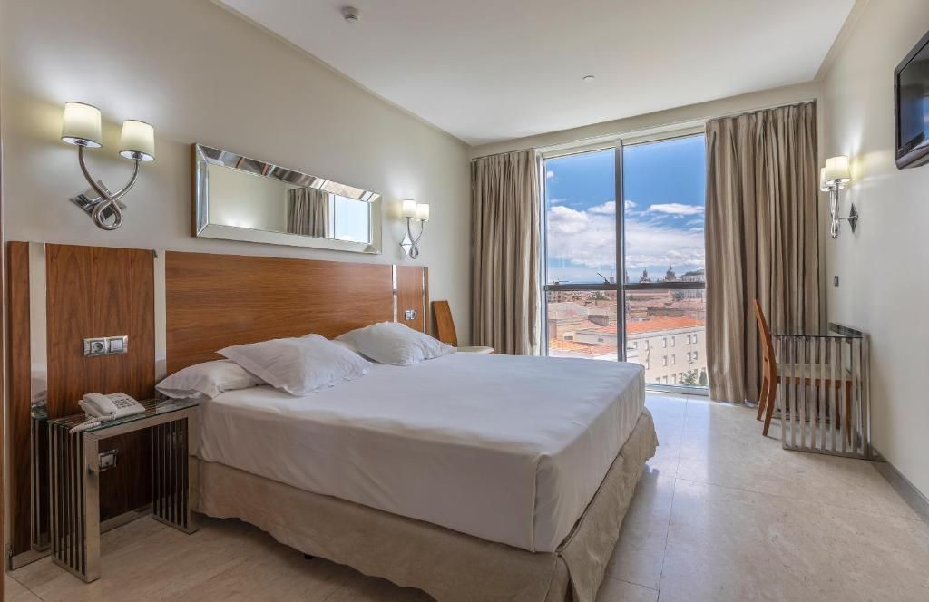 Habitación de hotel con una cama grande, un escritorio con teléfono, un espejo y vistas a la ciudad.