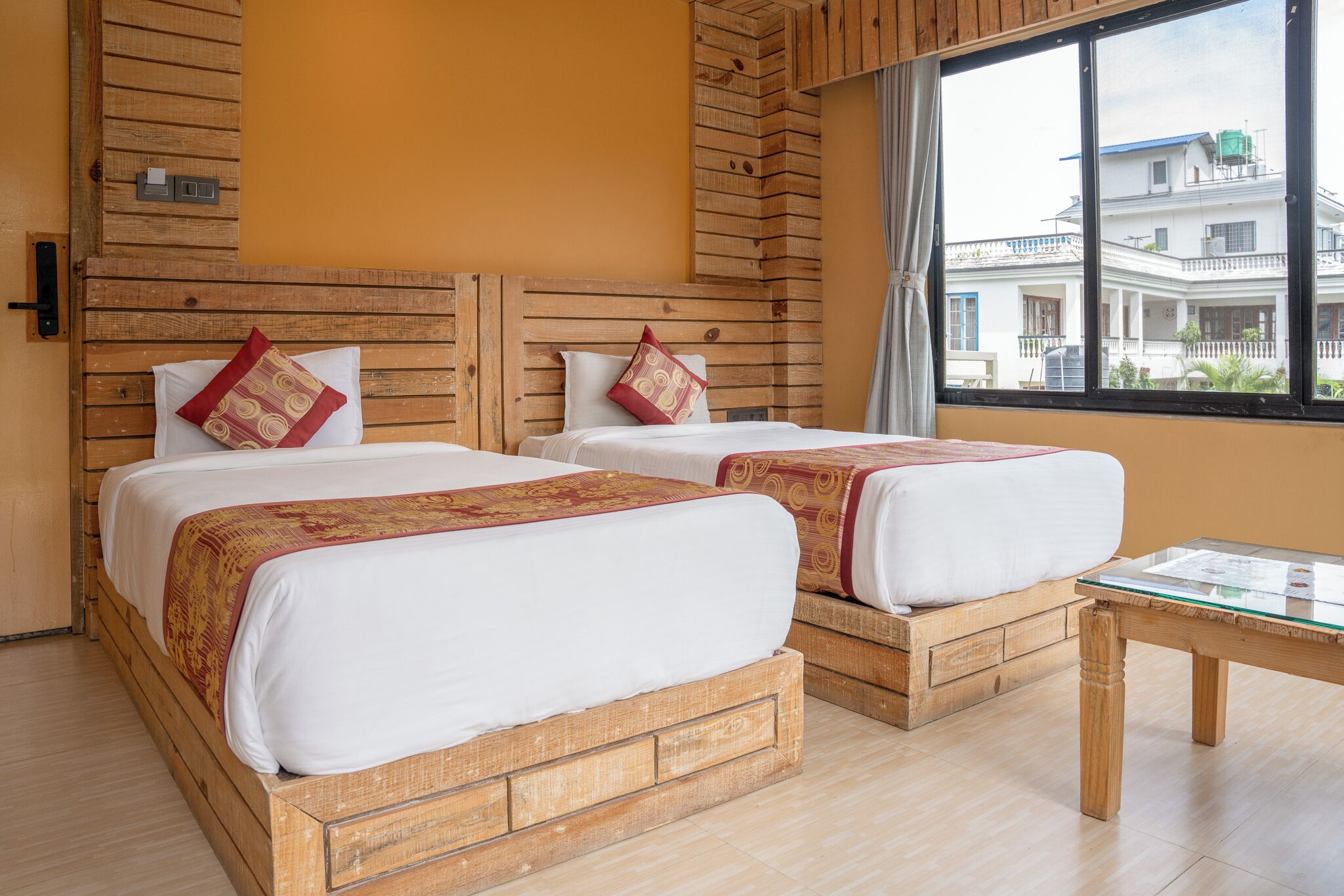 A room with two wooden beds, a glass table, and a view of a building outside.
