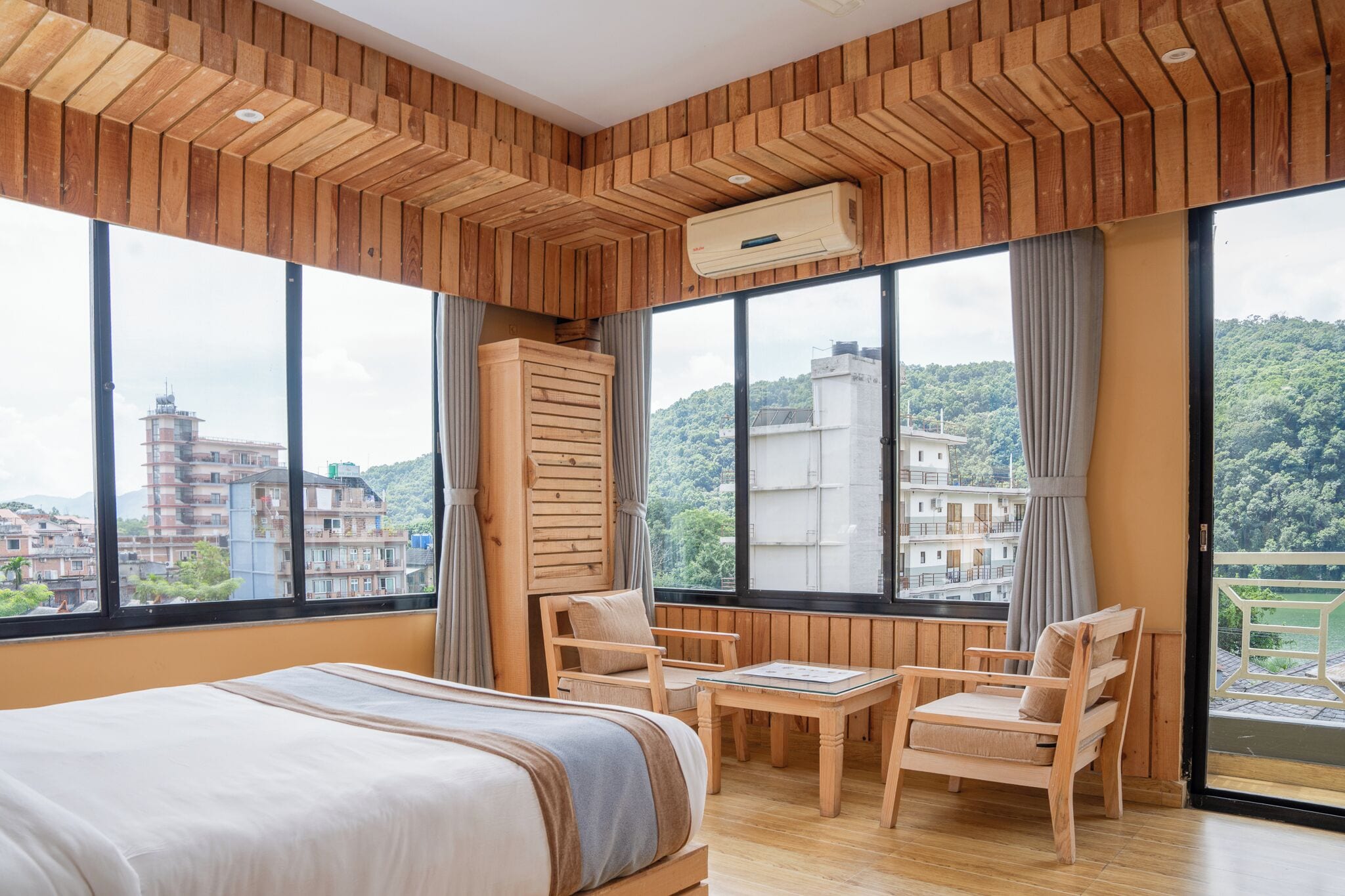 A hotel room with a bed, wooden furniture, large windows, and an air conditioning unit.