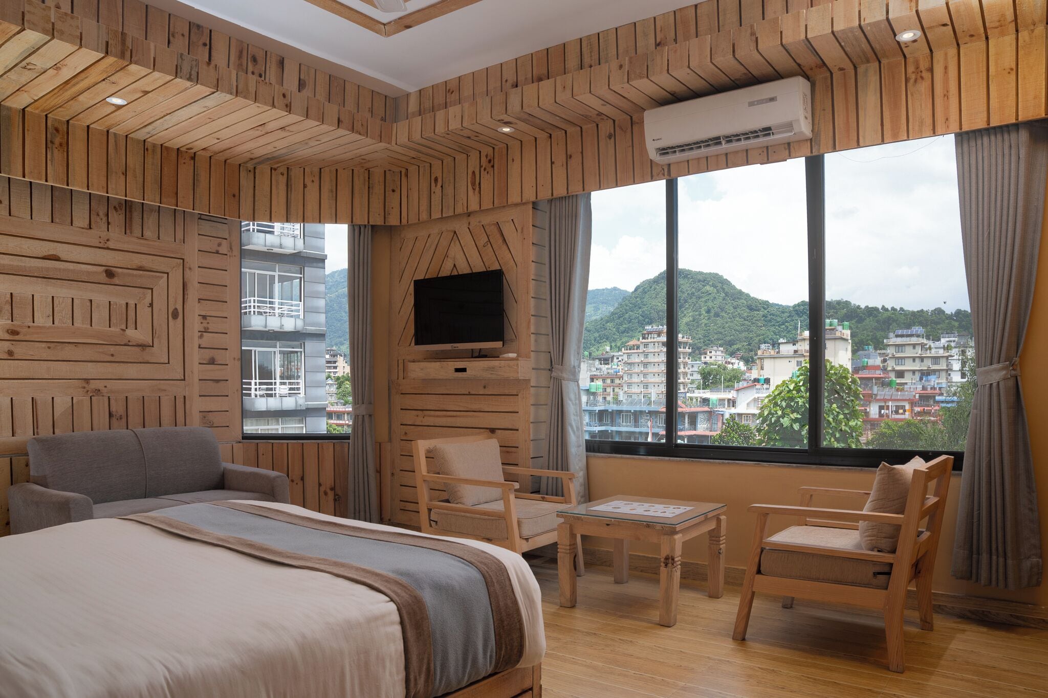 A hotel room with a large bed, two chairs, a small table, a TV mounted on a wooden wall, and a large window offering a city view.