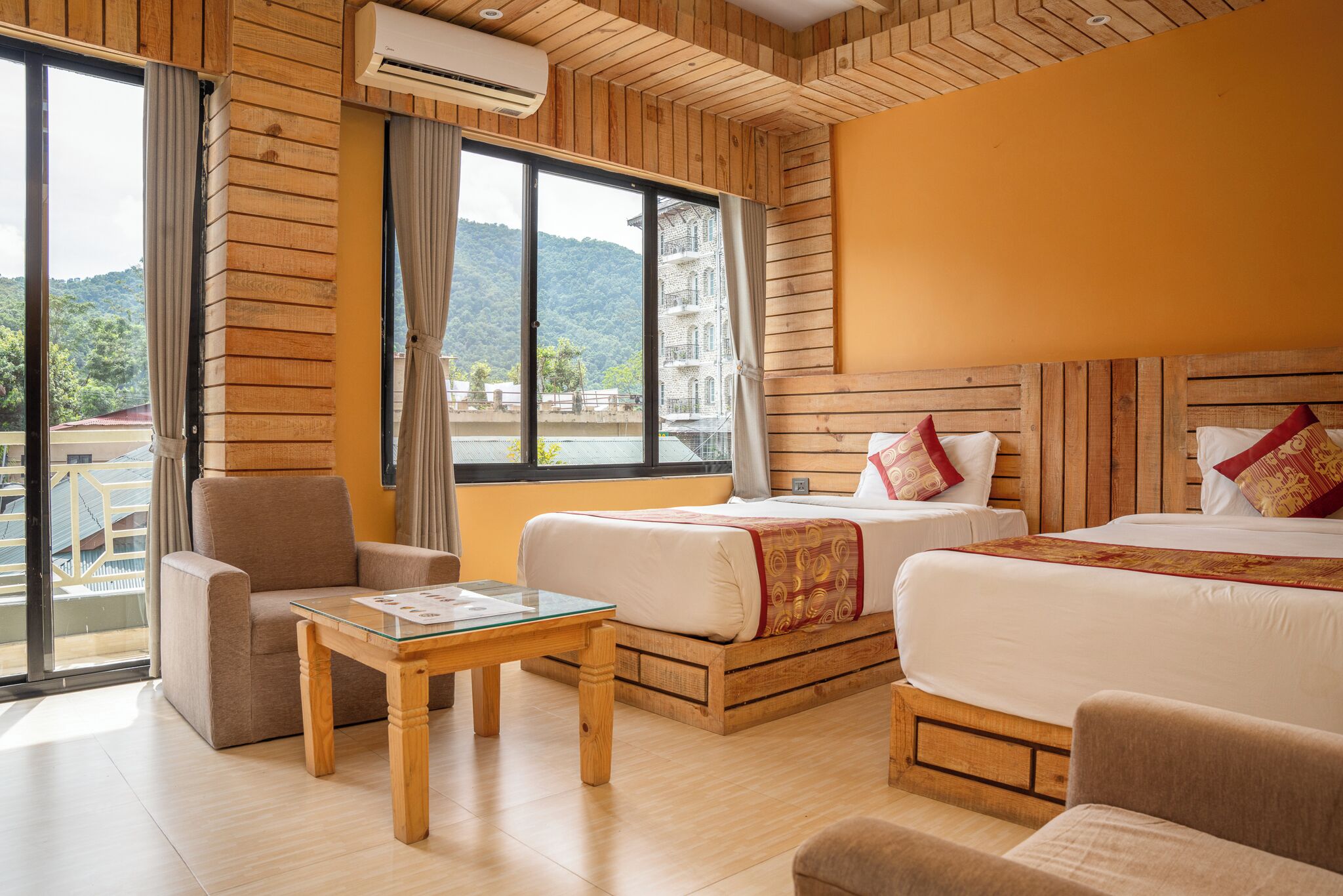 A hotel room with two beds, a sofa, a small table, and a large window offering a view of a building and hills.