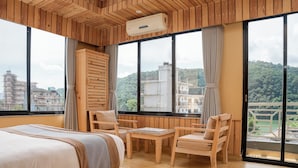Habitación de hotel con paneles de madera, cama, mesa y sillas. Cuenta con un ventanal con vistas a edificios y vegetación.