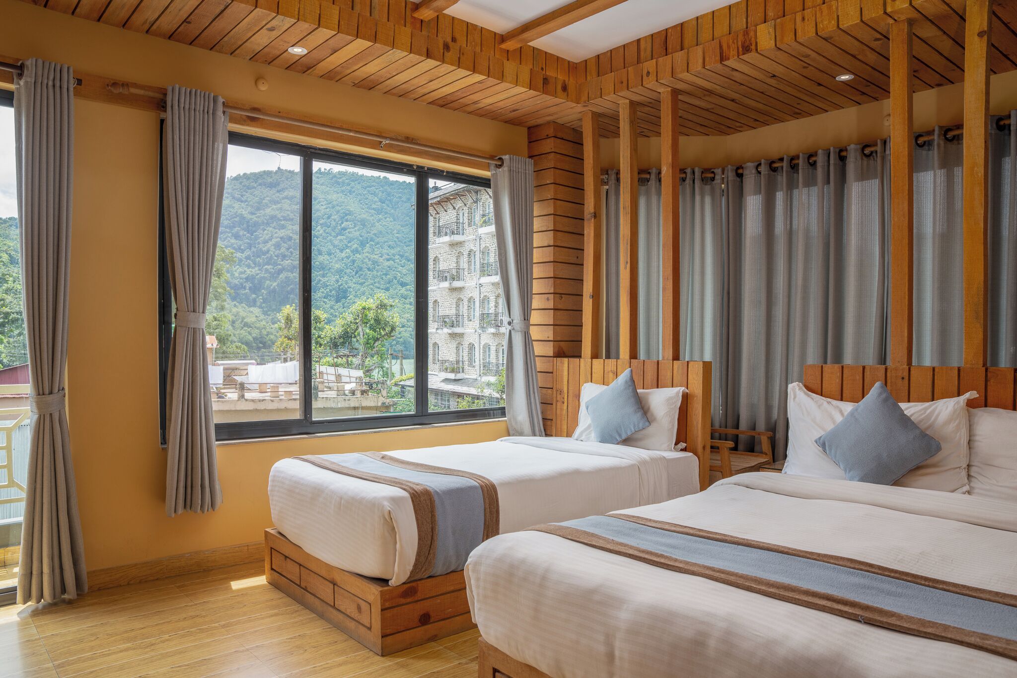 A hotel room with two beds, wooden headboards, large windows with curtains, and a view of a mountain and building.