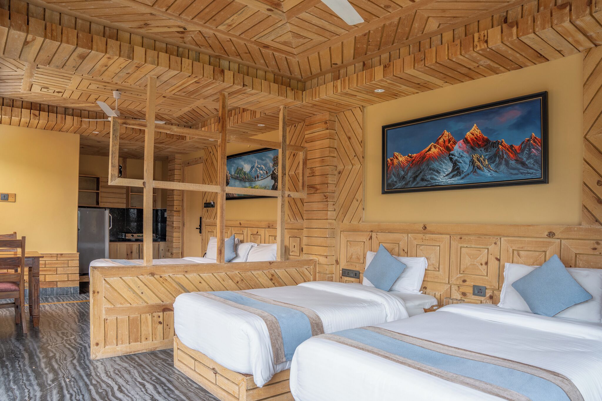 A hotel room with wooden paneling, two beds, a painting of mountains, and a kitchen area visible through an open door.