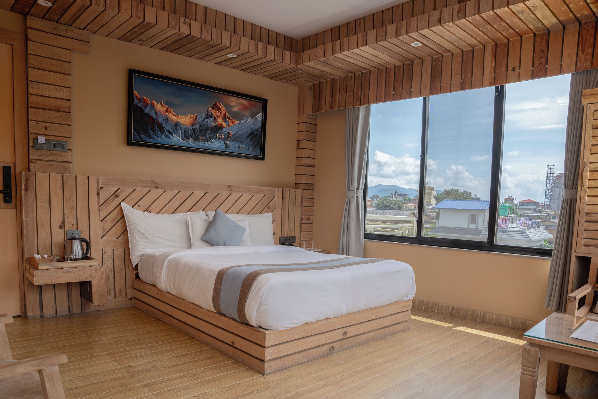 A modern bedroom with a wooden headboard, a large bed, a nightstand with a coffee maker, and a large window offering a city view.