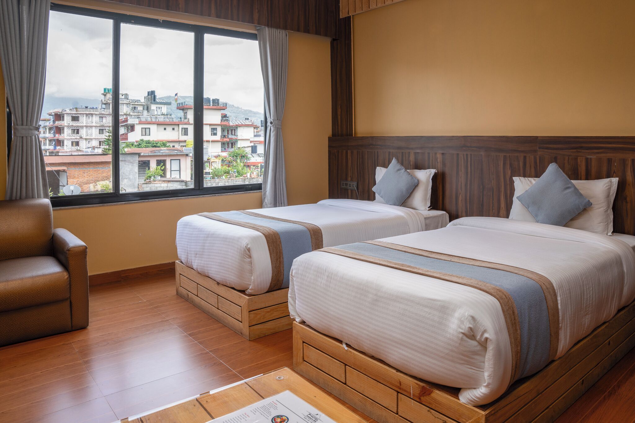 A hotel room with two beds, a wooden headboard, a window with curtains, a brown leather chair, and a wooden floor.