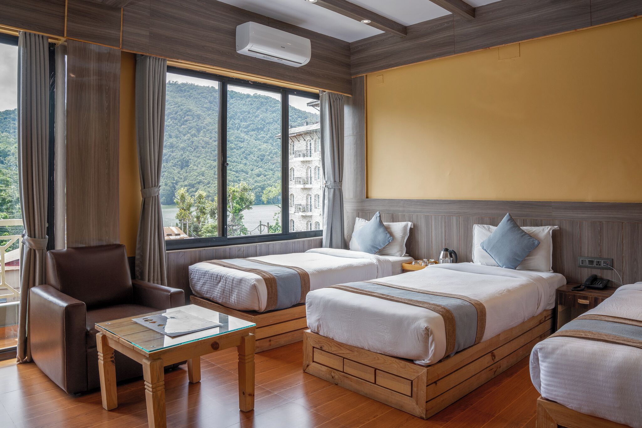 A hotel room with two beds, a sofa, a small table, and a large window offering a mountain view.