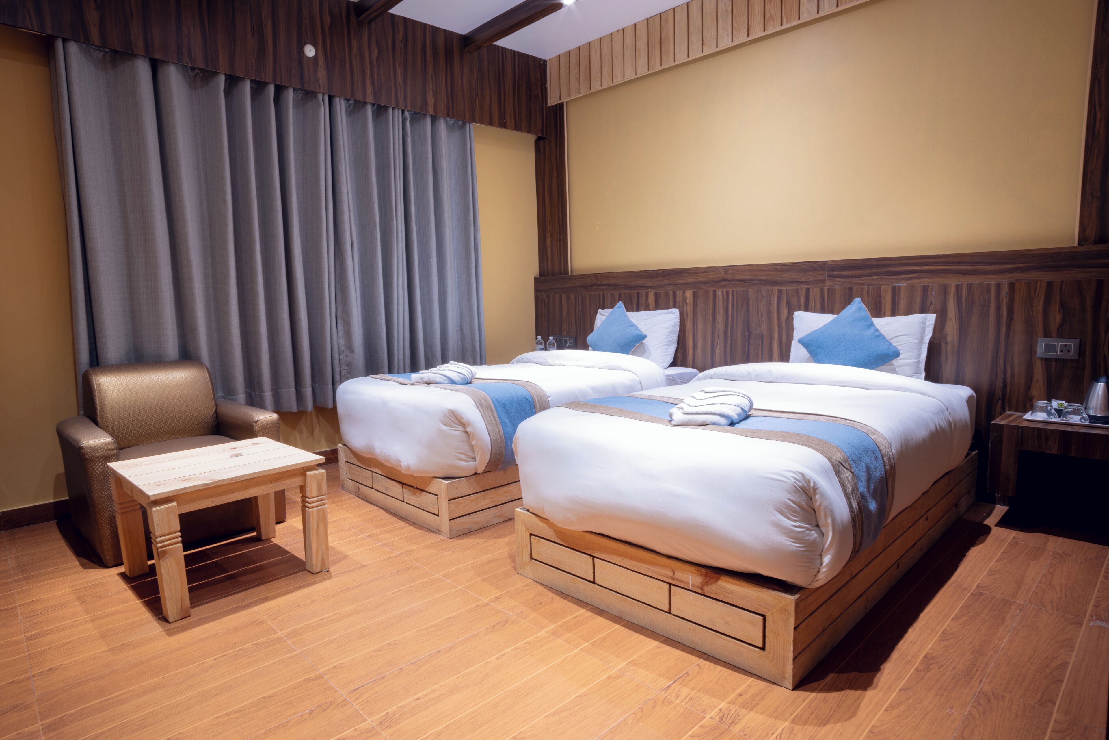 Habitación de hotel con dos camas, cabecera de madera, una mesita, una silla y una cortina.