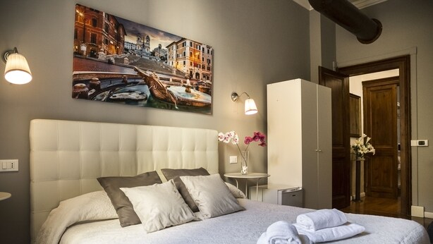 Una cama bien hecha con almohadas y una toalla doblada, un cuadro de un paisaje urbano en la pared, y una mesita con un jarrón de flores.