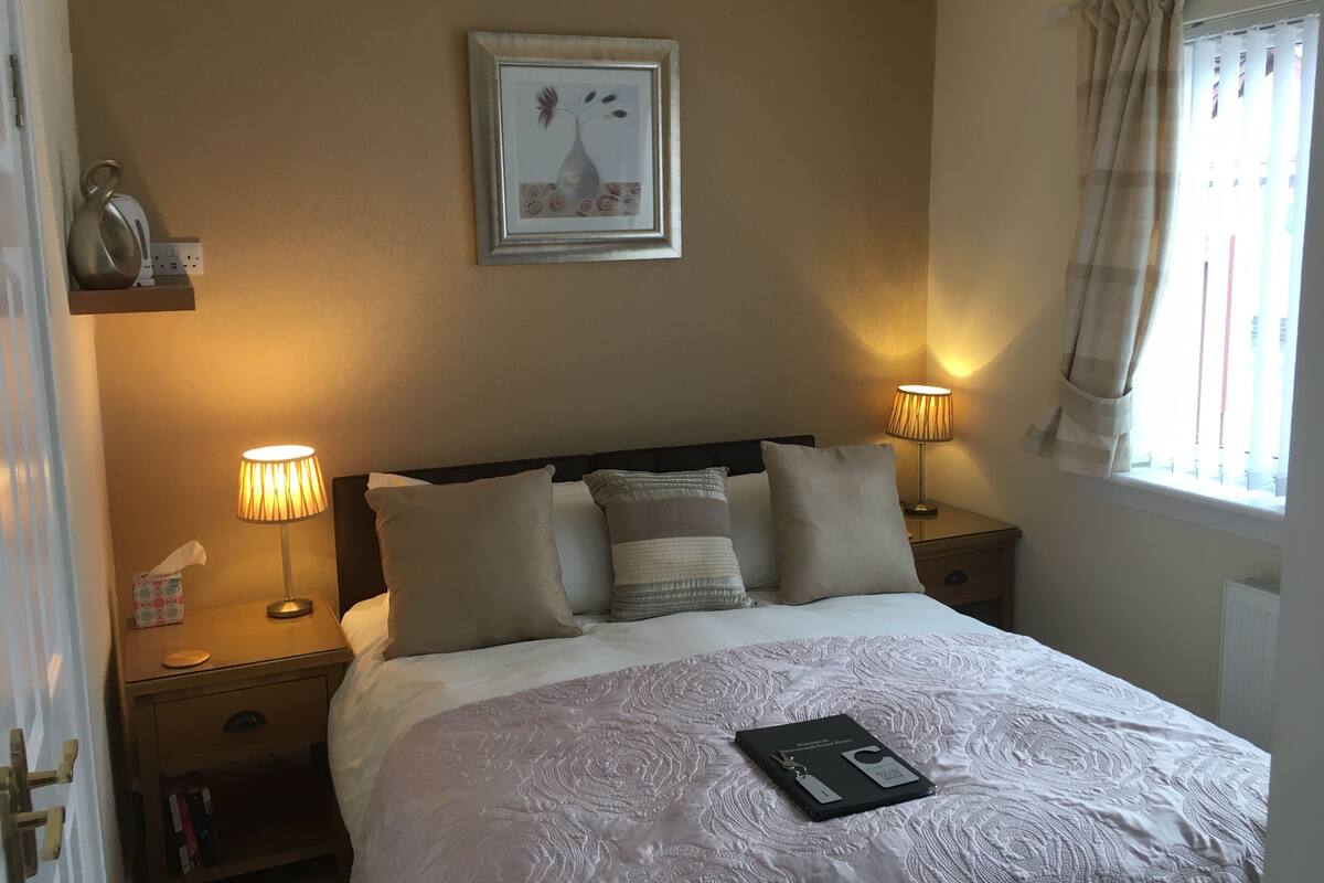 A neatly made bed with a white comforter and pillows, two bedside lamps, a framed artwork above the bed, and a window with curtains.