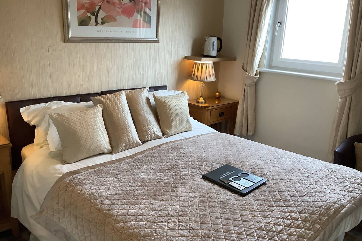A neatly made bed with a quilted cover, a framed picture on the wall, a bedside lamp, and a window with curtains.