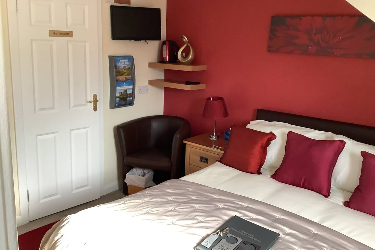 A hotel room with a bed, a chair, a nightstand, a TV, and a red wall.