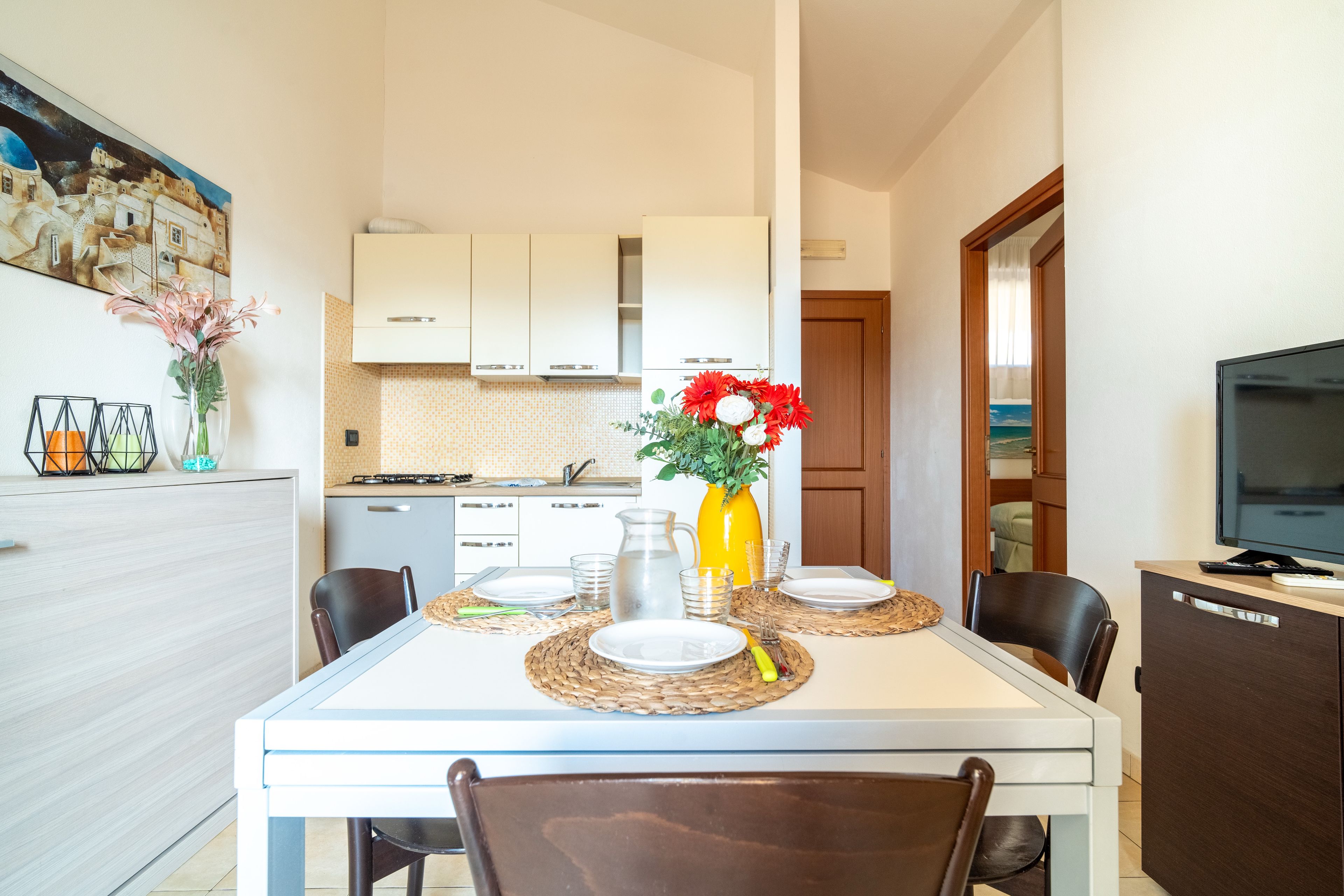 A dining area with a table set for two, a kitchen in the background, and a television on a cabinet.