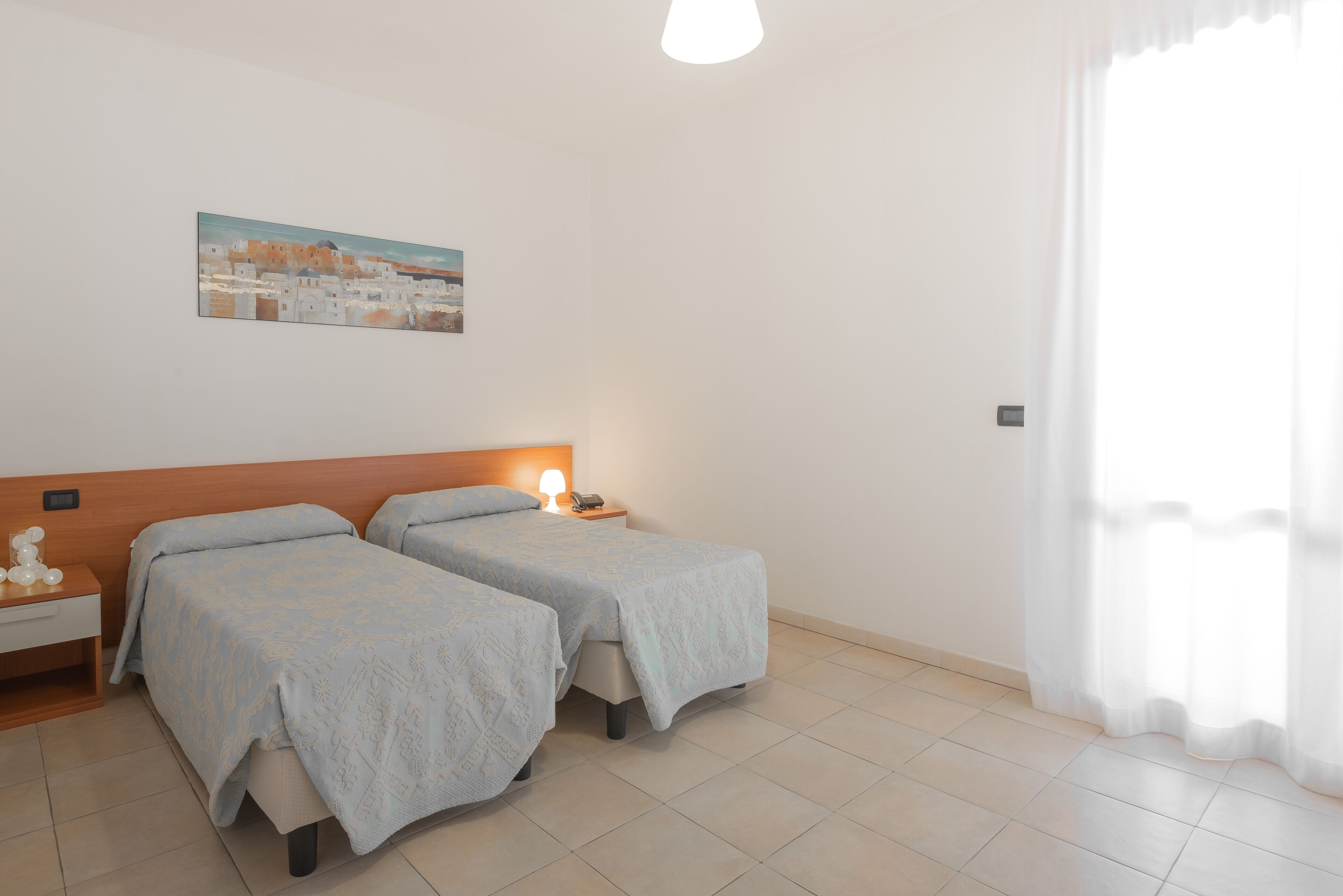 Una stanza con due letti, un comodino con lampada, un quadro appeso al muro e una finestra con tende.