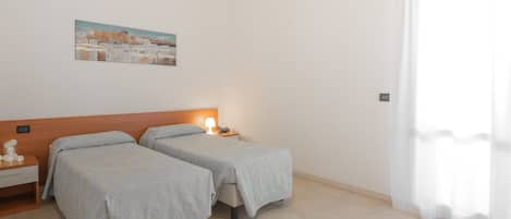 Una stanza con due letti, un comodino con lampada, un quadro appeso al muro e una finestra con tende.