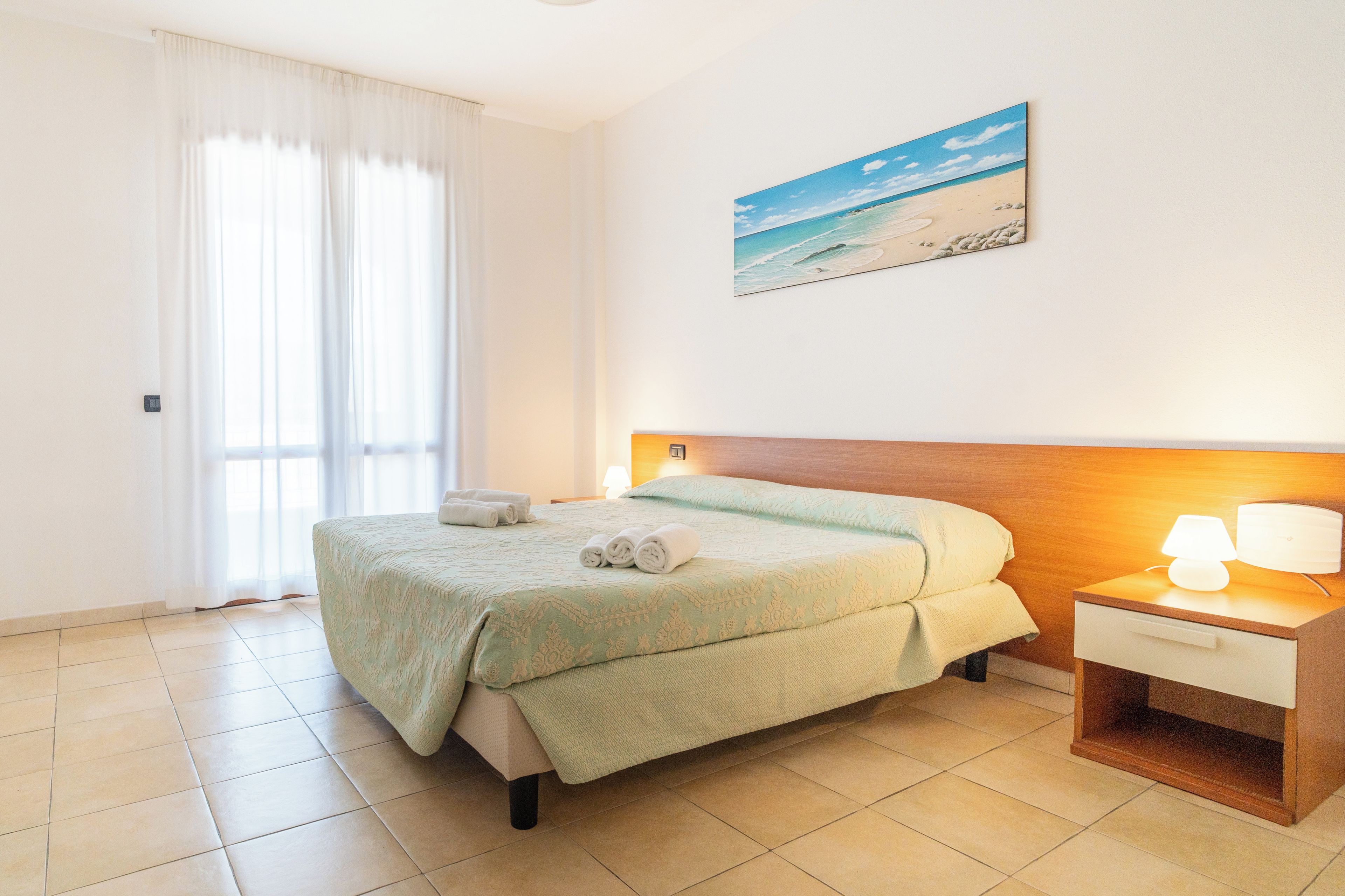 Habitación de hotel con cama, mesita de noche, lámpara, un cuadro de playa y una ventana con cortinas.