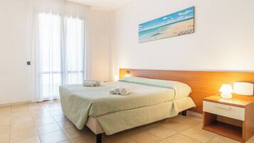 Habitación de hotel con cama, mesita de noche, lámpara, un cuadro de playa y una ventana con cortinas.