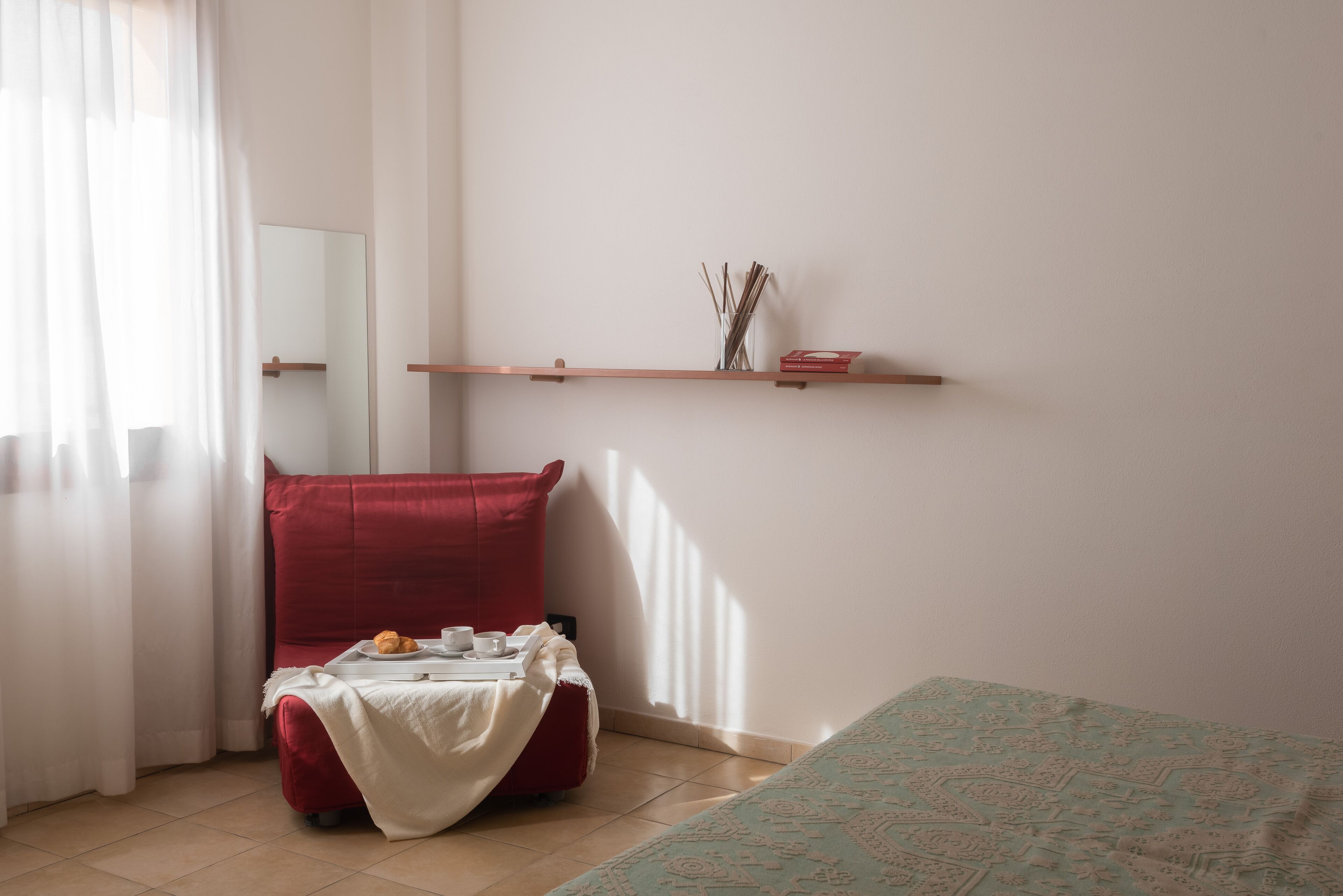 A room with a bed, a red chair, a small table with a tray of food, and a shelf with decorative items.