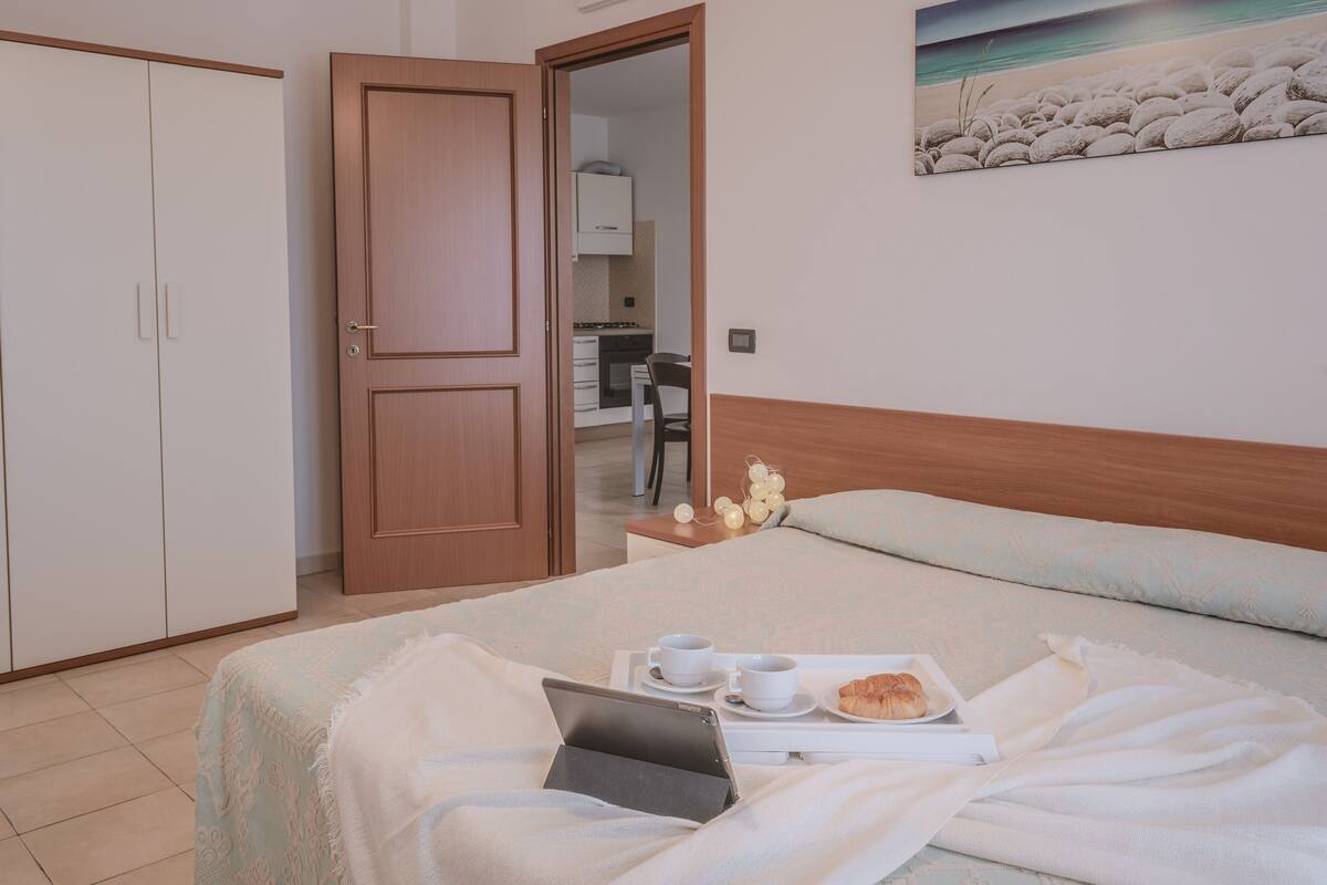 A neatly arranged bedroom with a bed, bedside table, and a view into the kitchen.
