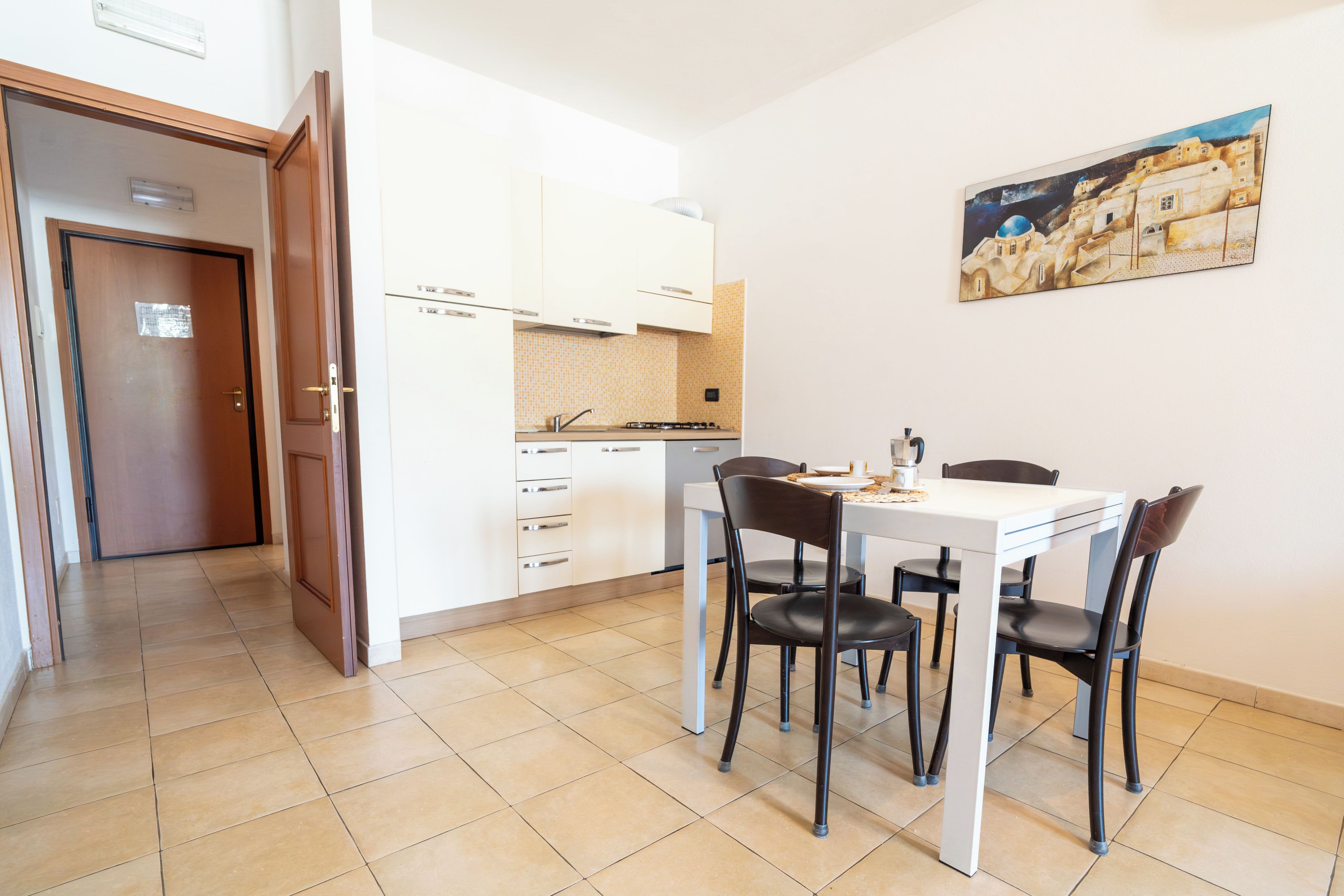 Une cuisine de taille réduite avec des meubles blancs, une table à manger et quatre chaises, ainsi qu’un tableau accroché au mur.