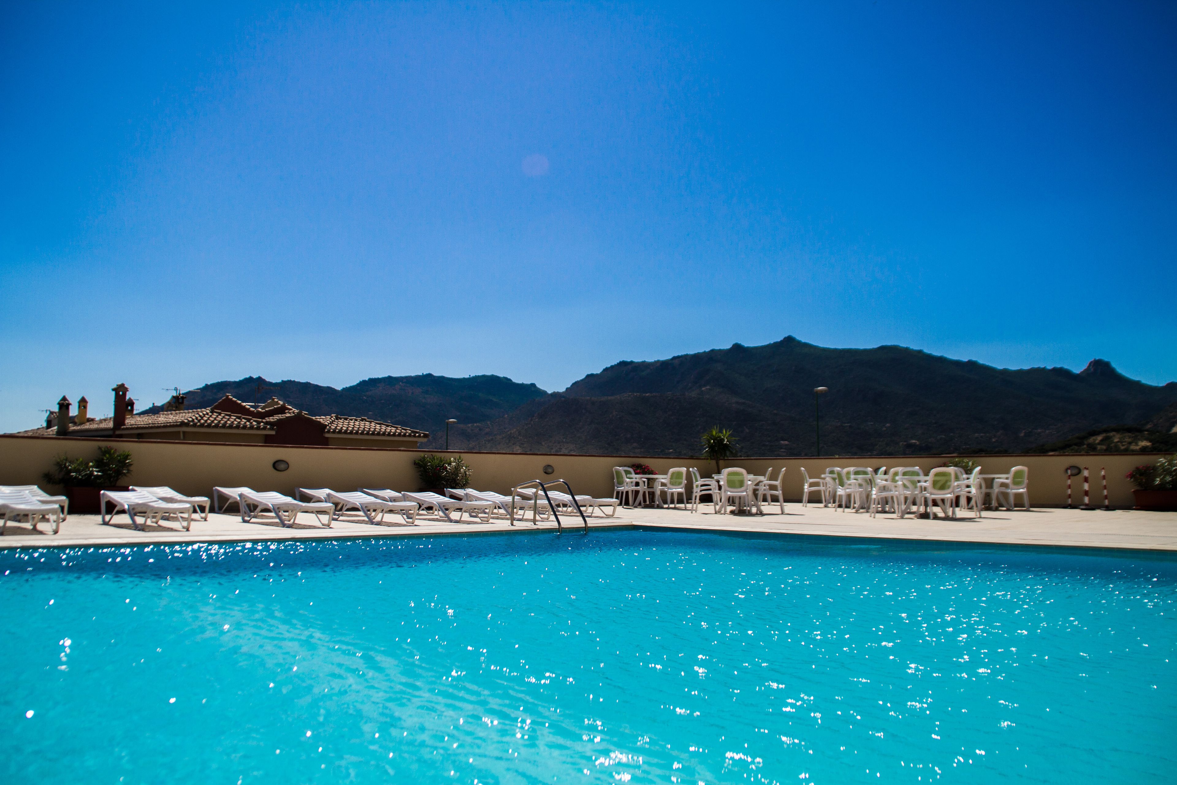 A swimming pool with lounge chairs and tables, surrounded by mountains.