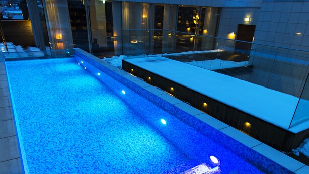 A swimming pool with blue lighting, surrounded by a modern building with large windows and a snow-covered roof. - PM & Vänner Hotel (Växjö)