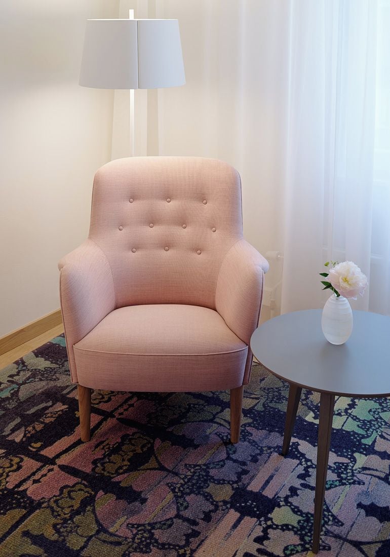A room with a pink upholstered armchair, a small round table with a vase of flowers, a patterned carpet, and a white lamp.