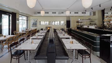 A spacious restaurant with long tables, a central bar, and a view of the city through large windows.